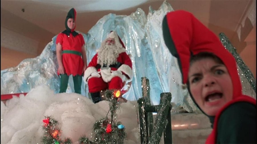 Santa Claus sits on an icy throne with two elves nearby; one elf is in the foreground with a surprised expression, while the other stands beside Santa. Holiday decorations and fake snow surround them.