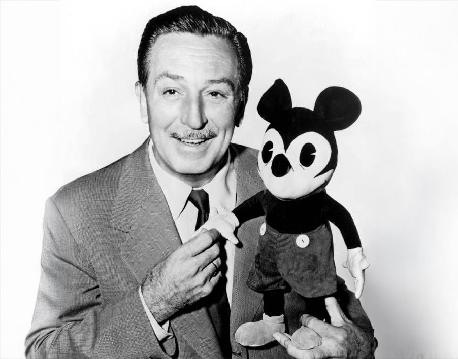A smiling man in a suit holds the hand of a vintage Mickey Mouse doll, standing against a plain white background.