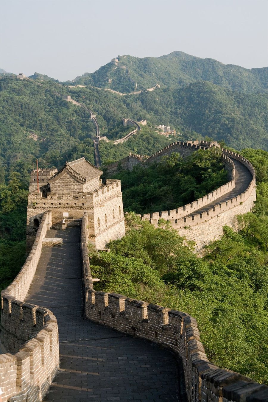 The Great Wall of China winds over green hills and mountains under a clear sky, showing stone pathways, watchtowers, and lush vegetation along the landscape.