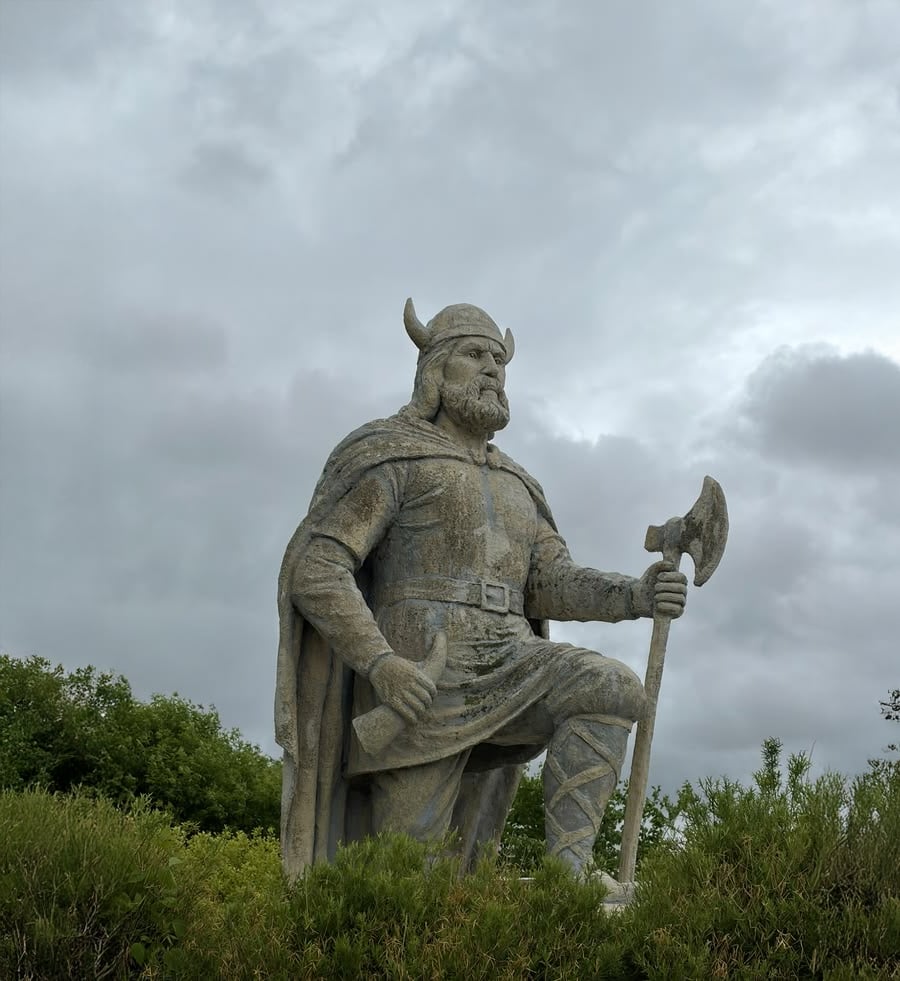 A stone statue of a Viking warrior stands outdoors, holding an axe and wearing a horned helmet and cape, with cloudy skies and green bushes in the background.