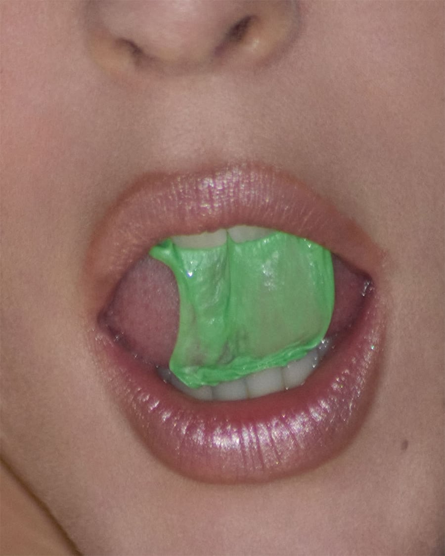 A close-up of a person’s mouth with glossy lips, slightly open, showing green chewing gum being stretched between their teeth and tongue.