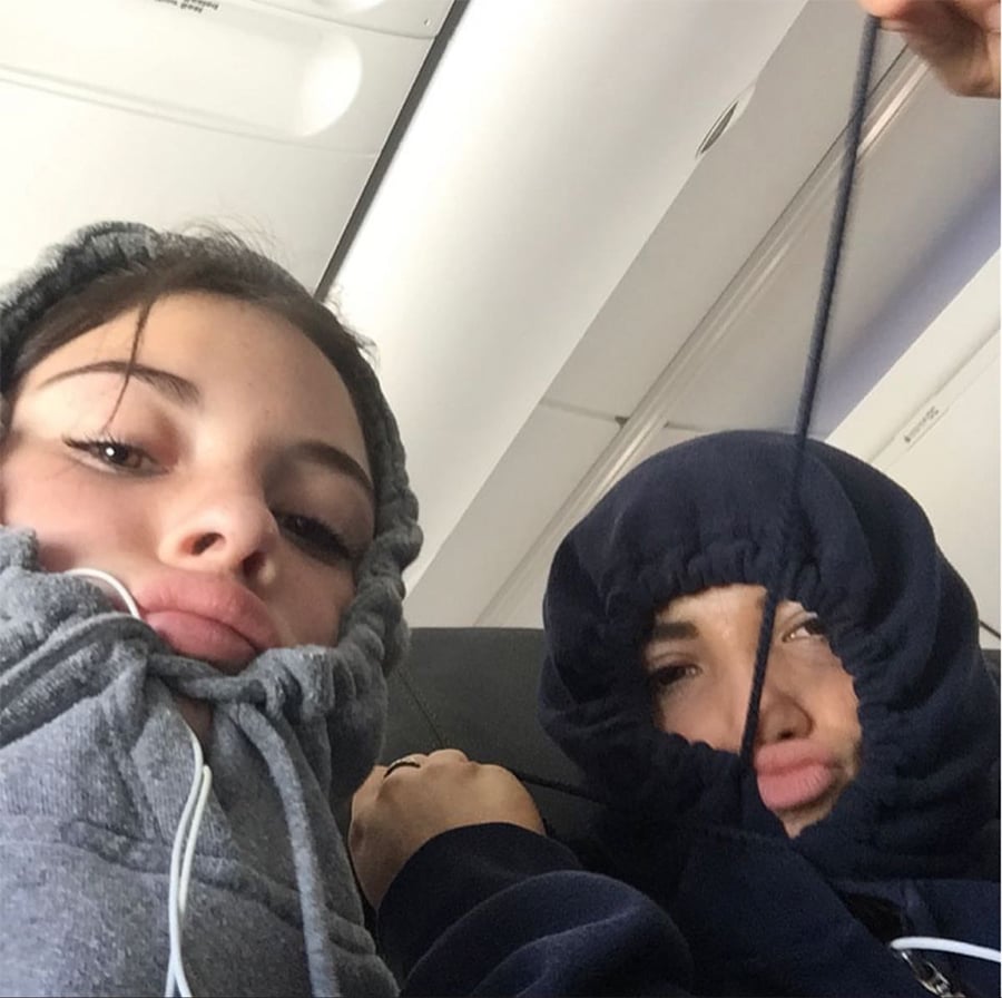 Two people sit on an airplane wearing hoodies pulled tightly around their faces, making playful expressions. One person has earphones, and their faces are partially obscured by the hoodie fabric.