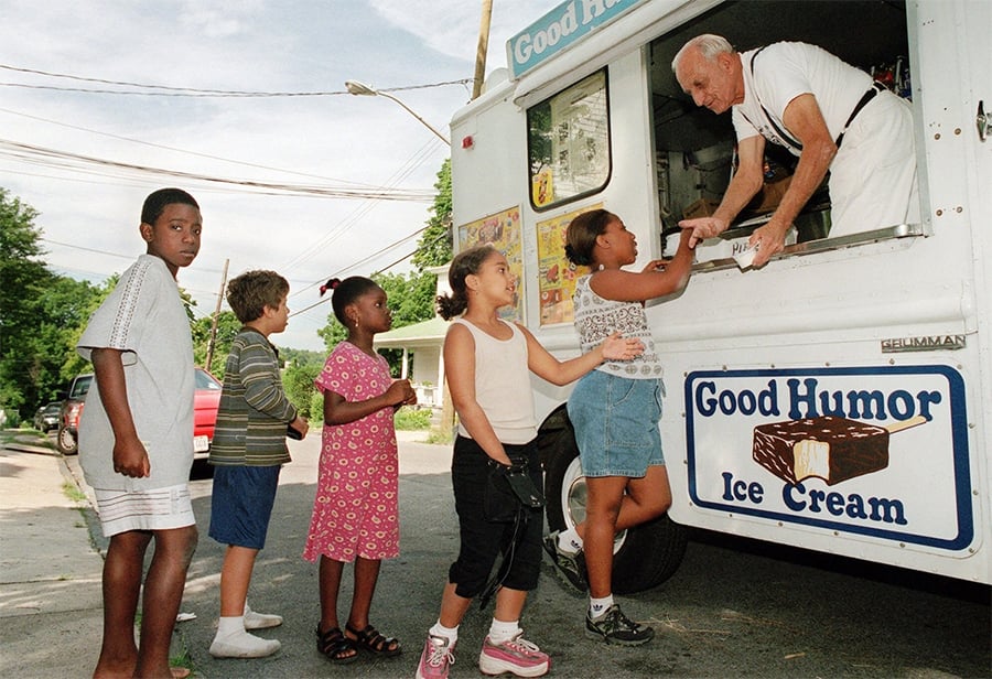 Five children stand in line at an ice cream truck, waiting to be served by an older man. The truck displays a Good Humor Ice Cream sign, and the scene takes place on a sunny neighborhood street.