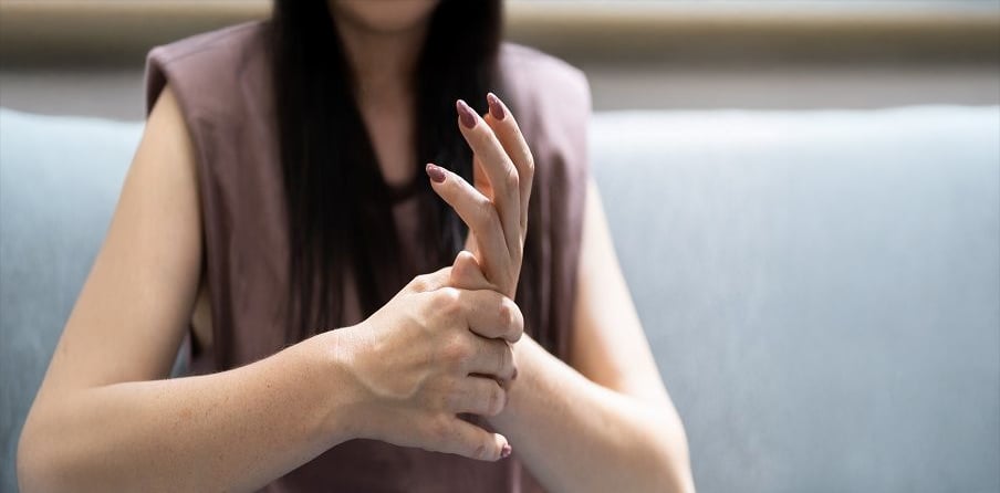 A woman sits on a sofa, holding her right wrist with her left hand, as if experiencing pain or discomfort in her wrist or hand. Her face is not visible.