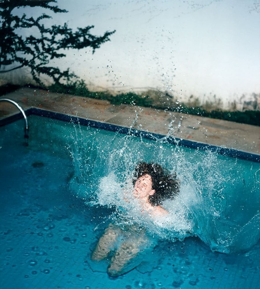 A person with curly hair is swimming in a pool, creating a big splash as water erupts around them. The bright blue water contrasts with the pool’s edge and surrounding greenery.