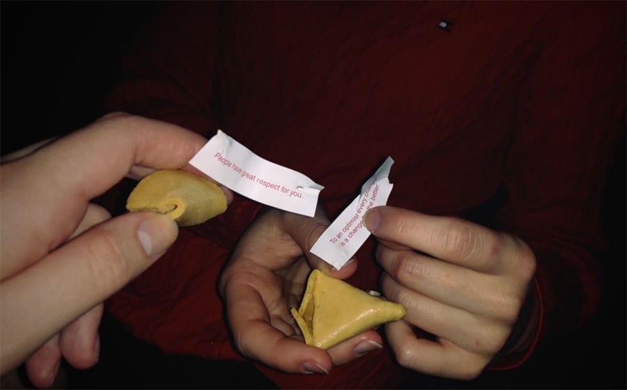 Two people hold broken fortune cookies and paper fortunes. One fortune reads, “People have great respect for you.” Both individuals wear red clothing.