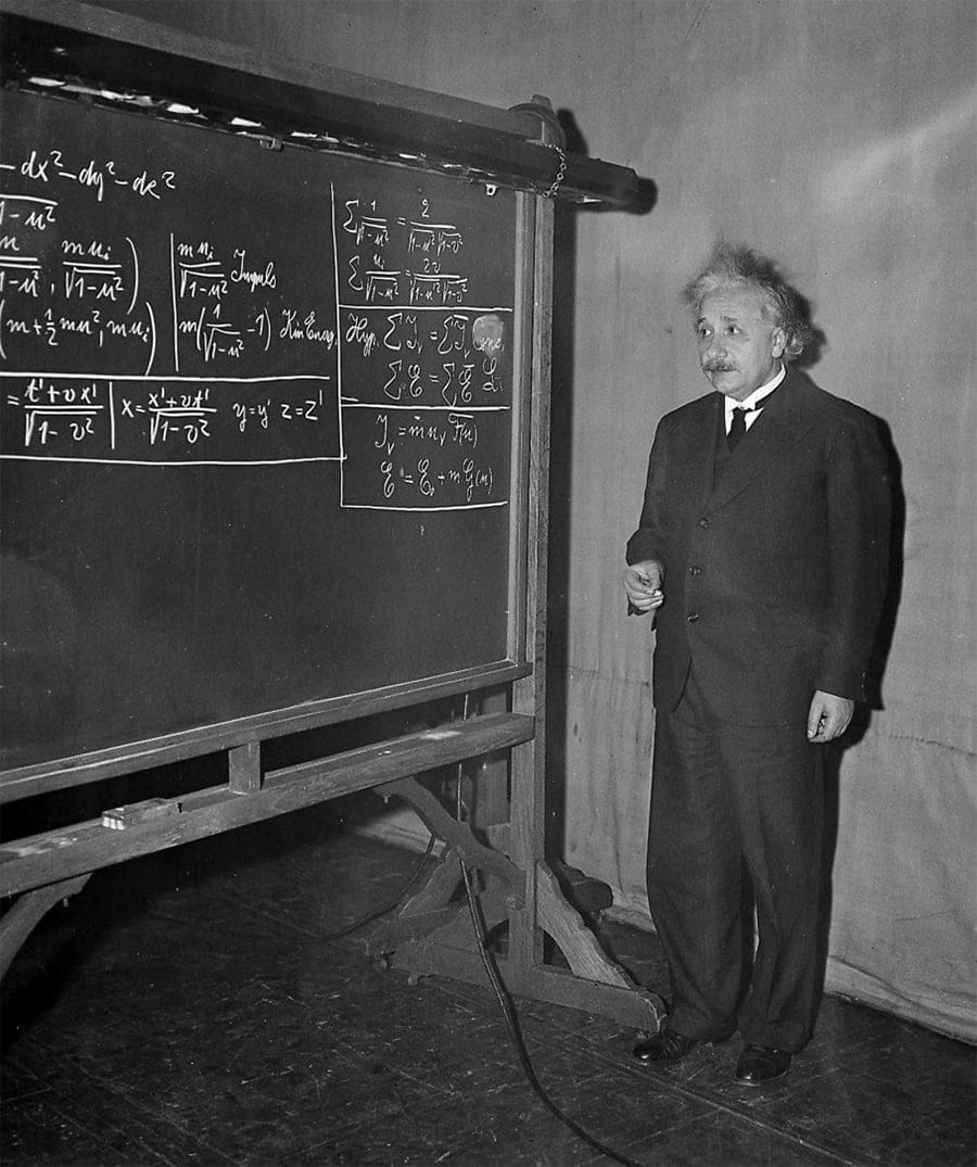 A man in a suit stands next to a large chalkboard covered with complex mathematical equations, appearing to lecture or explain concepts. The setting looks like a classroom or lecture hall.