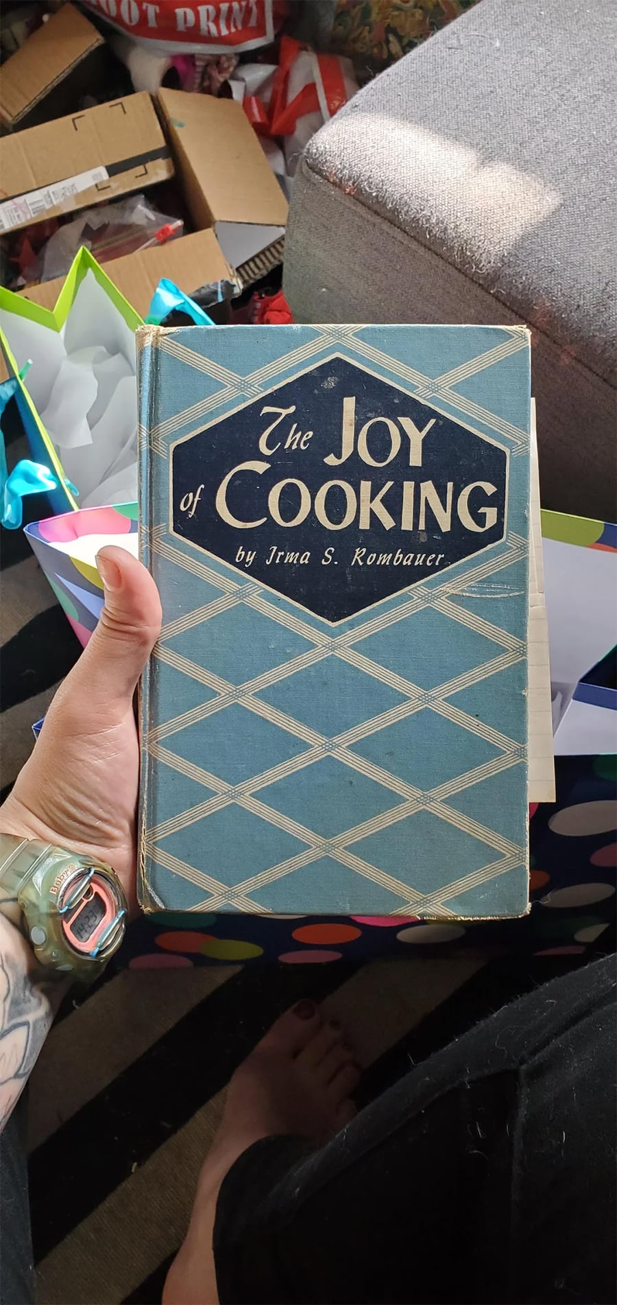 A hand holds a vintage copy of “The Joy of Cooking” by Irma S. Rombauer. The book has a blue geometric cover. Background includes gift bags, boxes, and part of a gray couch.