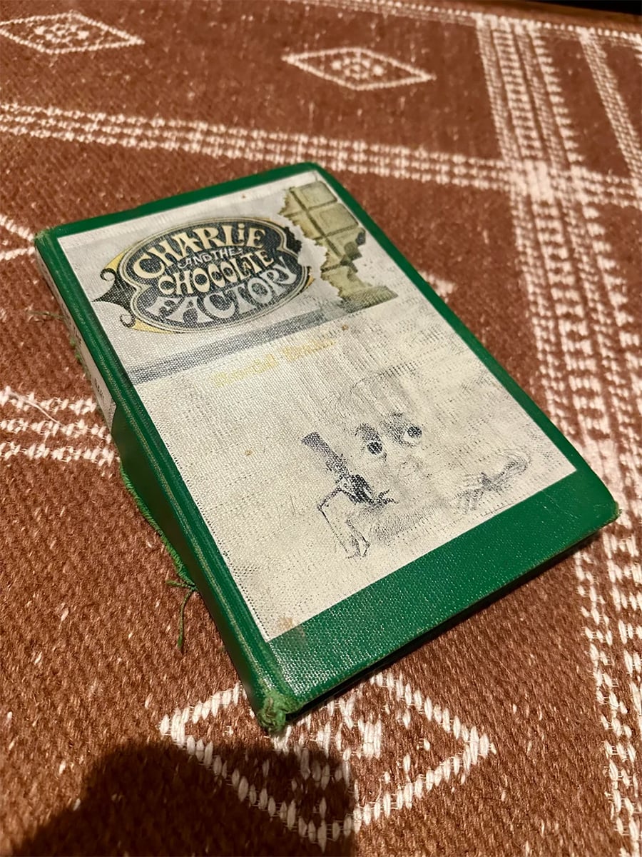 A worn, green hardcover book of “Charlie and the Chocolate Factory” rests on a patterned brown surface. The cover shows fading and visible damage along the spine.