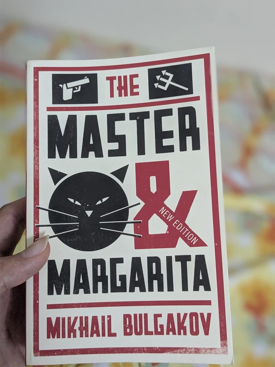 A hand holds the book “The Master & Margarita” by Mikhail Bulgakov, featuring bold black and red graphics of a cat, a gun, and a sickle and hammer on the cover.