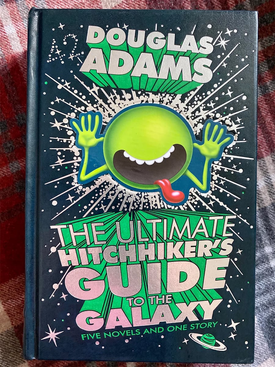Book cover of “The Ultimate Hitchhiker’s Guide to the Galaxy” by Douglas Adams, featuring a green, smiling alien with its tongue out and hands raised, surrounded by stars on a dark background.