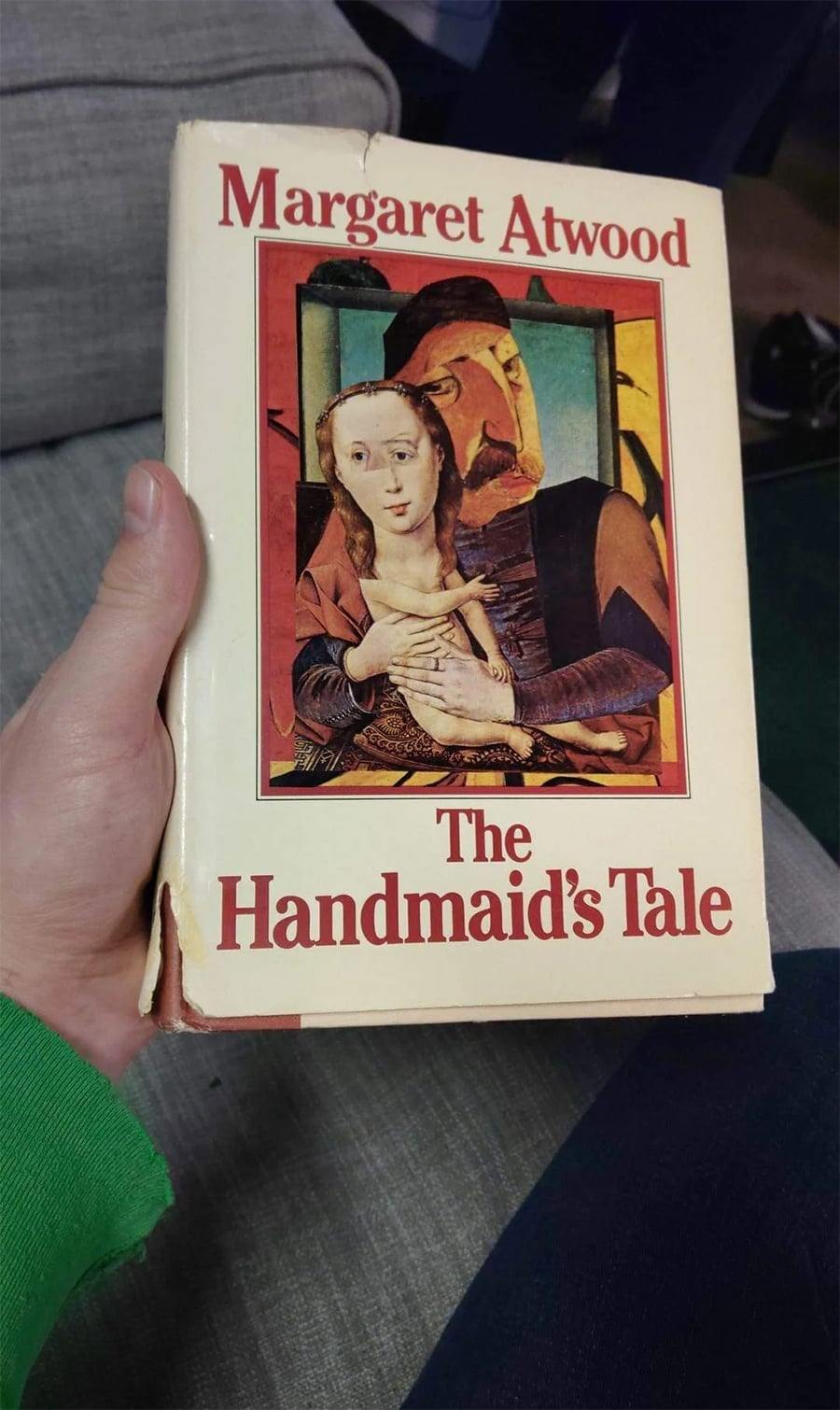 A hand holds a worn copy of “The Handmaid’s Tale” by Margaret Atwood, featuring a cover illustration of two figures in a stylized, classical art style.