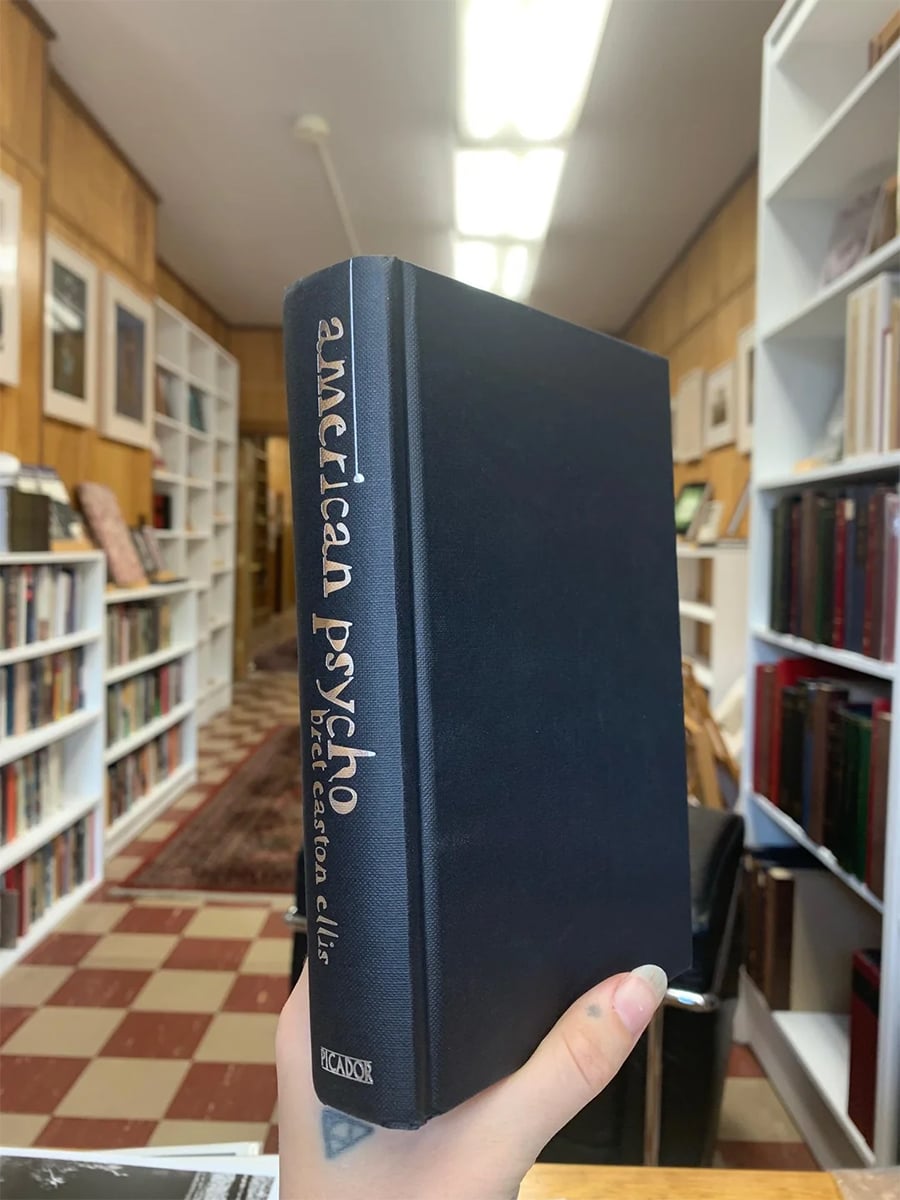 A hand holds a hardcover copy of “American Psycho” by Bret Easton Ellis in a bookstore with shelves of books and framed art on the walls.