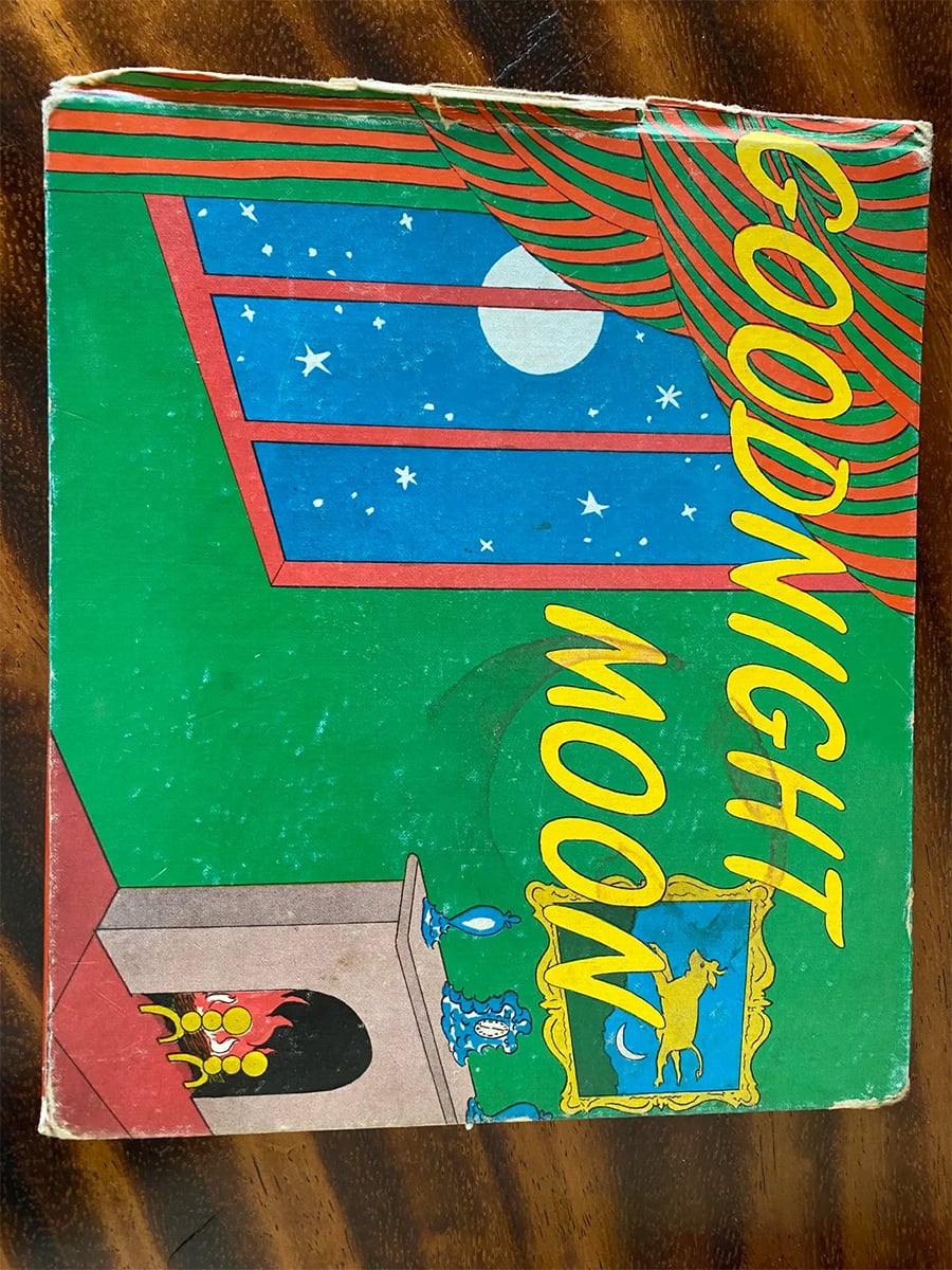 The cover of the book “Goodnight Moon” shows a room with a fireplace, blue balloons, a window with a night sky, and the title in large yellow letters.