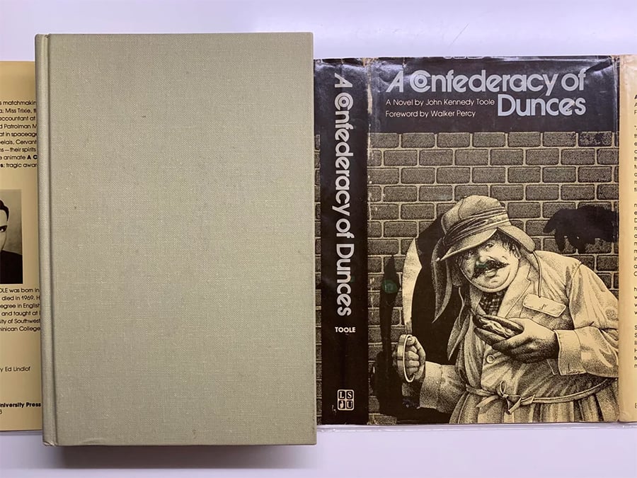 The book “A Confederacy of Dunces” by John Kennedy Toole is shown with its dust jacket featuring an illustrated man in a brown coat and hat, standing in front of a brick wall. The cover and spine are visible.