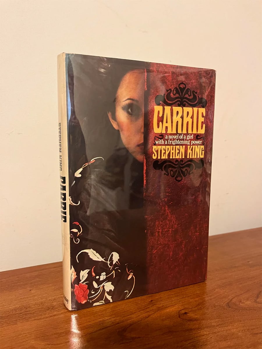 A hardcover copy of Stephen King’s “Carrie” stands on a wooden surface. The cover shows a woman’s face partly veiled in shadow, with the title and author’s name in bold yellow and white text.