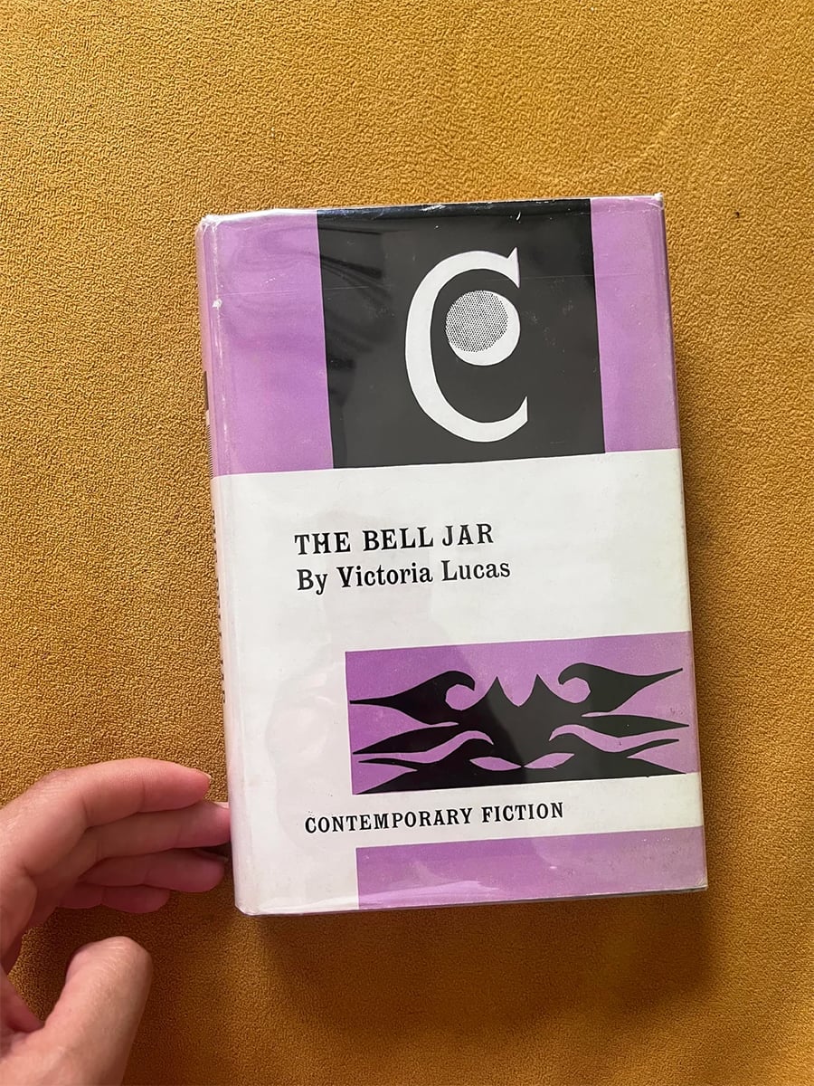 A hand holds a copy of “The Bell Jar” by Victoria Lucas, featuring a purple, black, and white cover with abstract design and the label “Contemporary Fiction.”