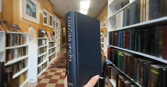 A hand holds the book “American Psycho” by Bret Easton Ellis in the aisle of a cozy bookstore lined with shelves of colorful books and framed artwork.
