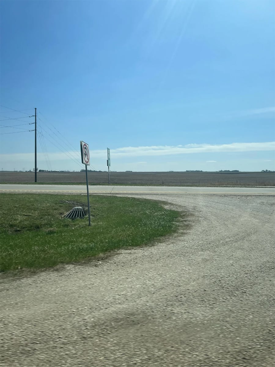 A gravel road curves toward a paved highway next to a vast, flat field under a clear blue sky. A “No U-Turn” sign stands on a grassy patch near a metal storm drain grate, and a highway exit sign is visible in the distance.