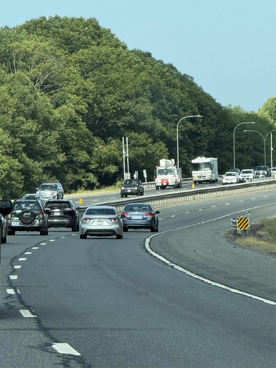 Cars drive on a divided highway bordered by trees, with vehicles traveling in both directions. The sky is clear, and the road curves to the right in the foreground.