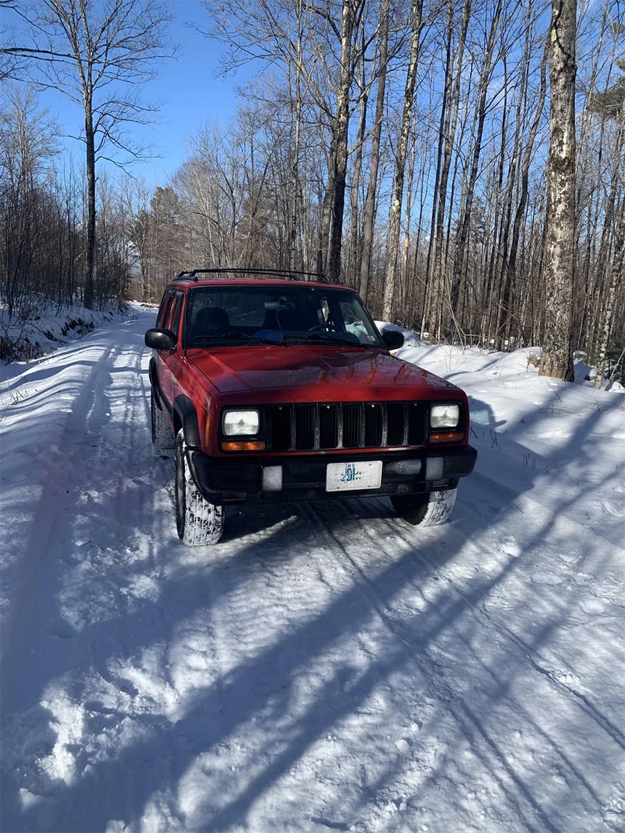 A red SUV drives on a snowy, tree-lined road under a clear blue sky. Tire tracks are visible in the snow, and bare trees flank both sides of the road.