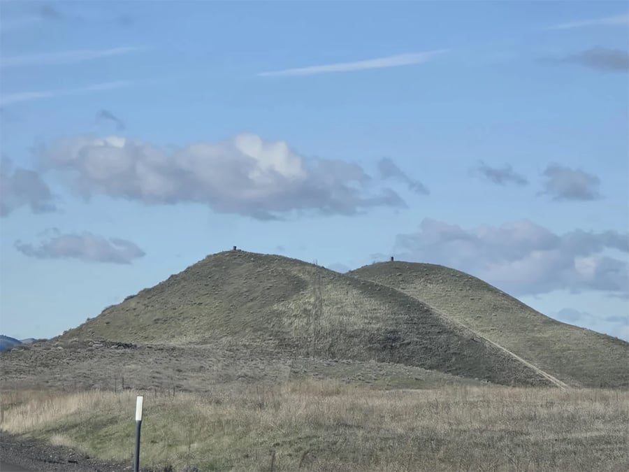 Two grassy, rounded hills under a blue sky with scattered clouds. A faint path runs between the hills, and a small white signpost is visible in the foreground.