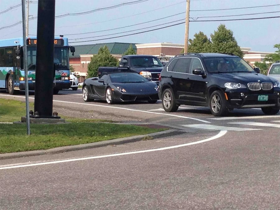 A black Lamborghini convertible waits in traffic behind a black SUV at an intersection, with a public bus and other vehicles nearby. Trees and a building are visible in the background.