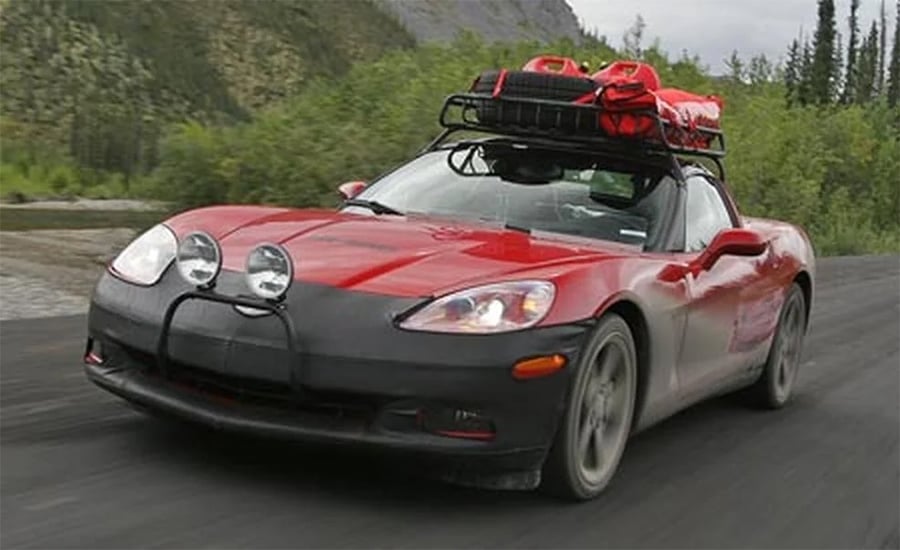 A red Chevrolet Corvette with off-road lights and a roof rack loaded with gear drives on a dirt road surrounded by trees and hills.