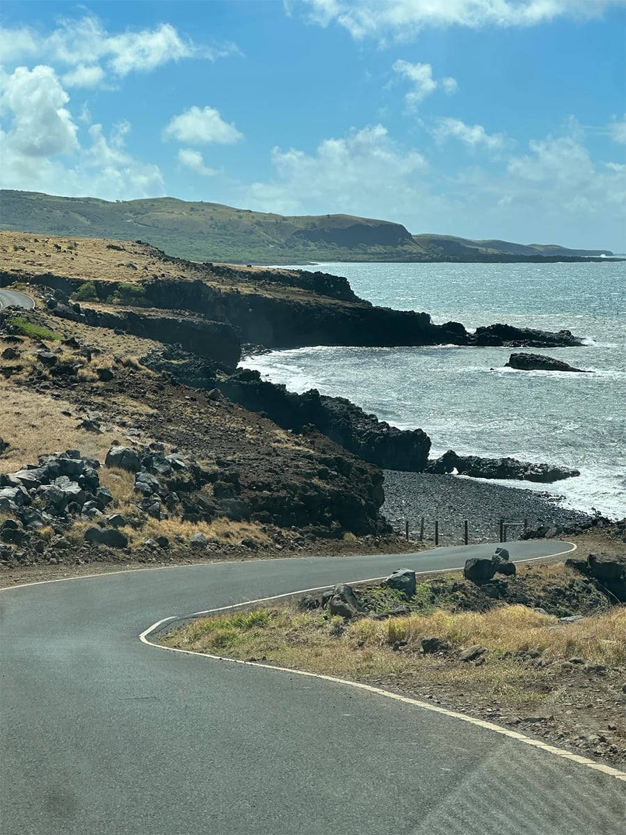 A winding coastal road curves along rocky cliffs beside the ocean, with waves crashing on dark rocks and rolling green hills in the background under a partly cloudy sky.