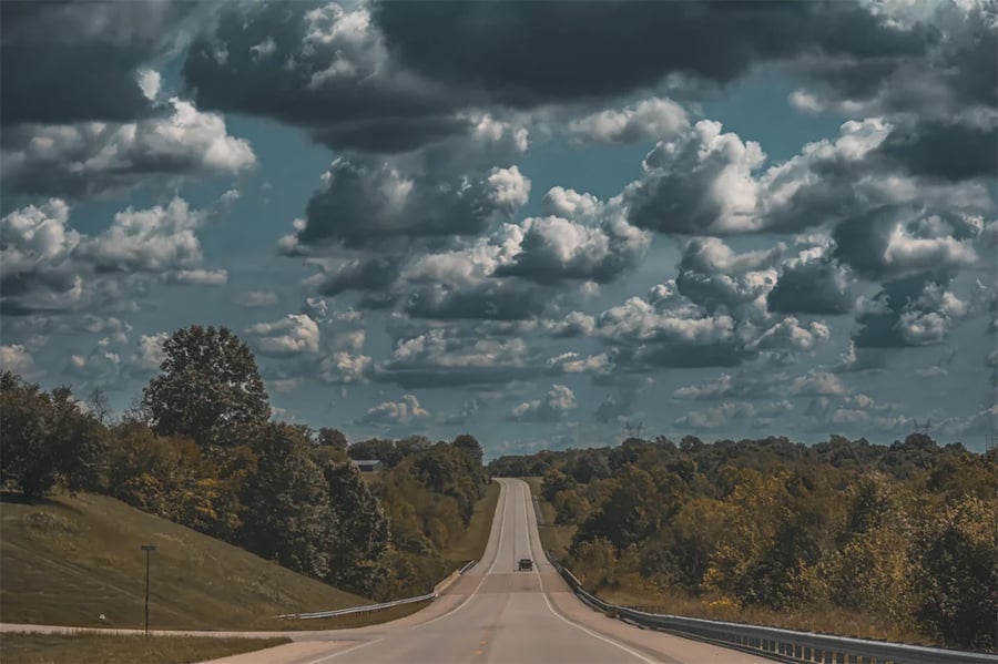 A car drives down a rural highway surrounded by trees under a dramatic, cloudy sky with patches of sunlight.