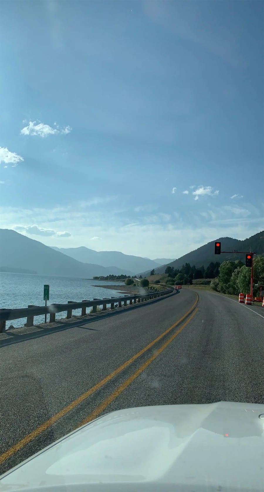 A winding road curves along a lake with mountains in the distance. Red traffic lights and orange cones appear on the right side under a clear blue sky.