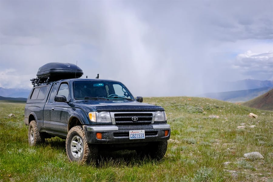 A black Toyota pickup truck with a roof cargo box is parked on grassy terrain under a cloudy sky, with hills and a misty or rainy area visible in the background.