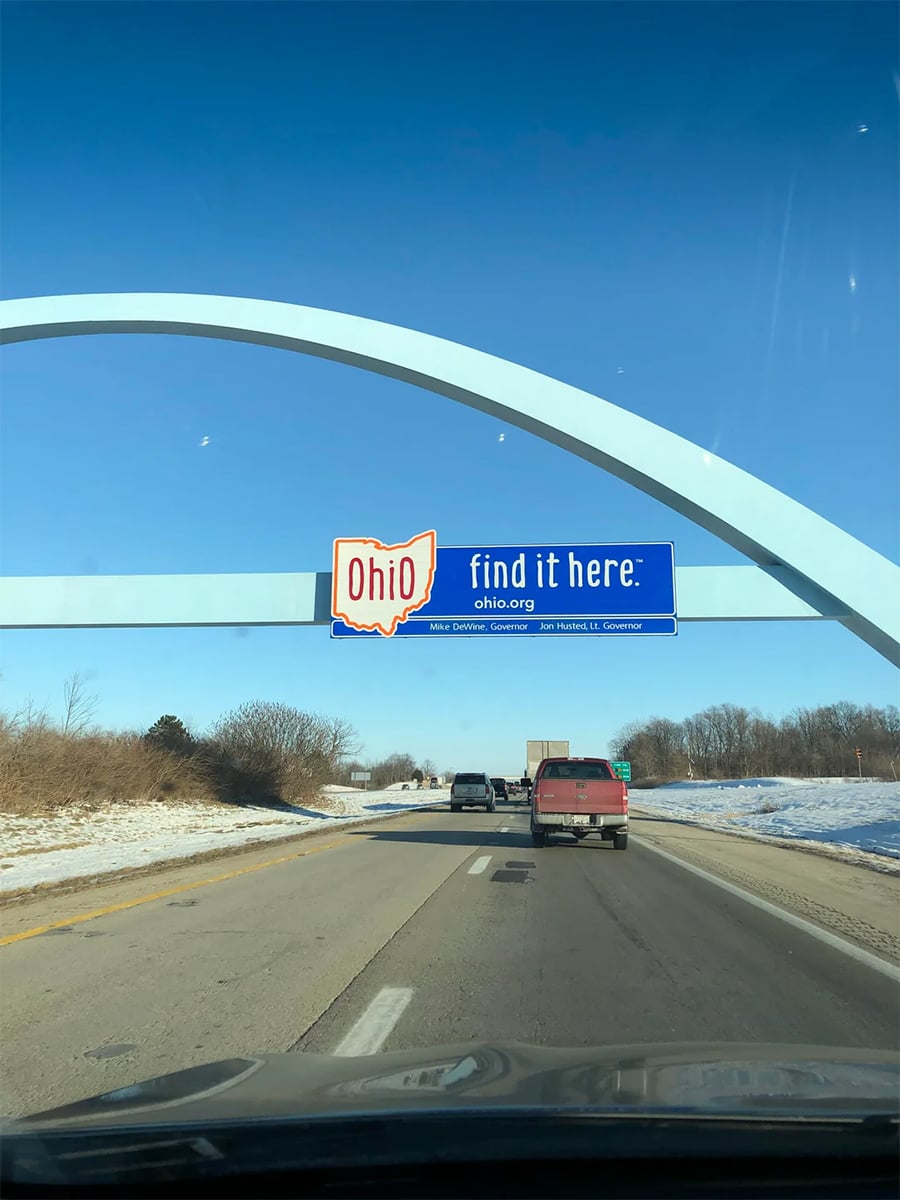 Cars drive under a blue arch with a sign reading “Ohio find it here.” on a sunny winter day, with snow along the sides of the highway and clear skies overhead.
