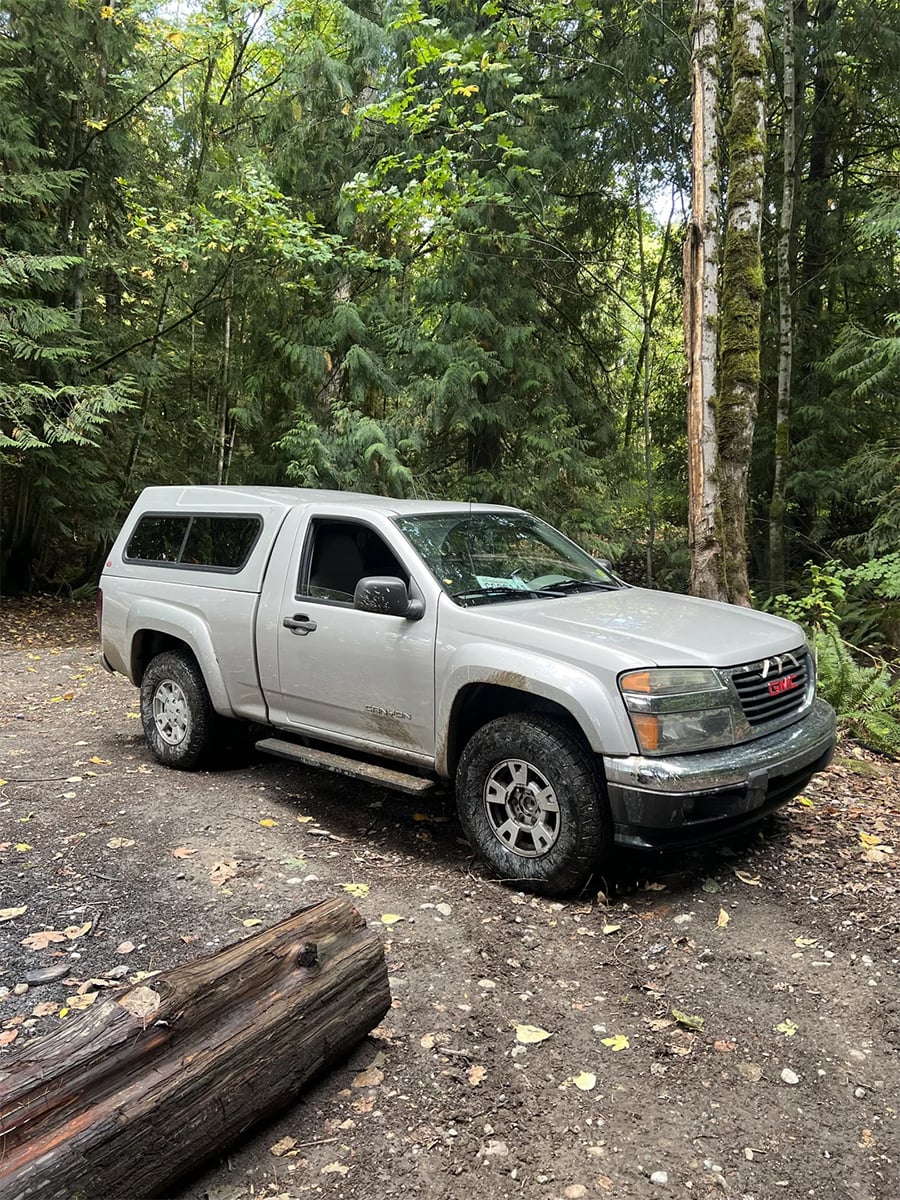 A silver GMC pickup truck with a matching camper shell is parked on a dirt clearing surrounded by lush green trees in a forest. A log lies in the foreground.