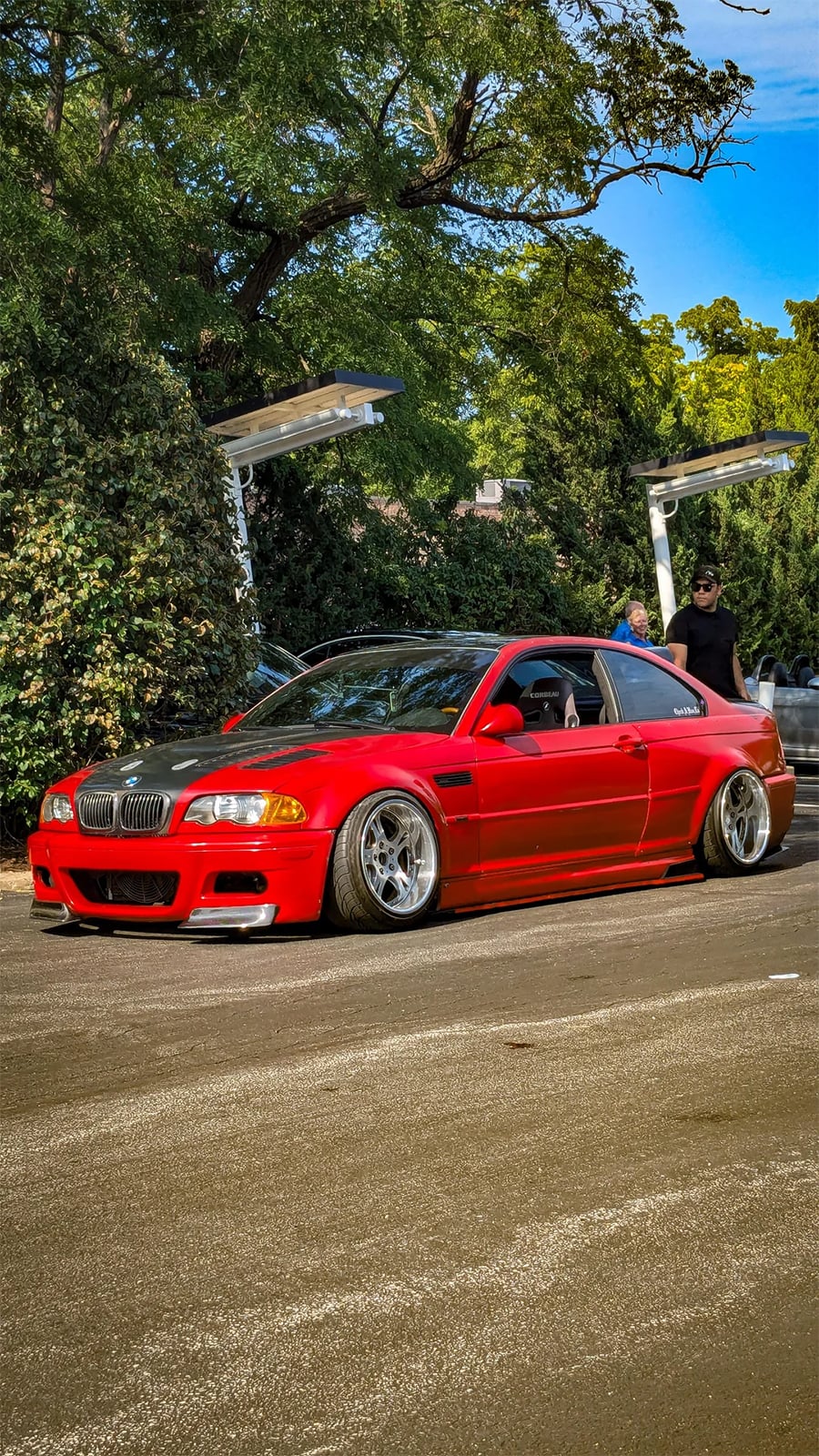 A red modified BMW coupe with lowered suspension is parked outdoors. Two people stand nearby under solar-powered lights, with trees and greenery in the background on a sunny day.