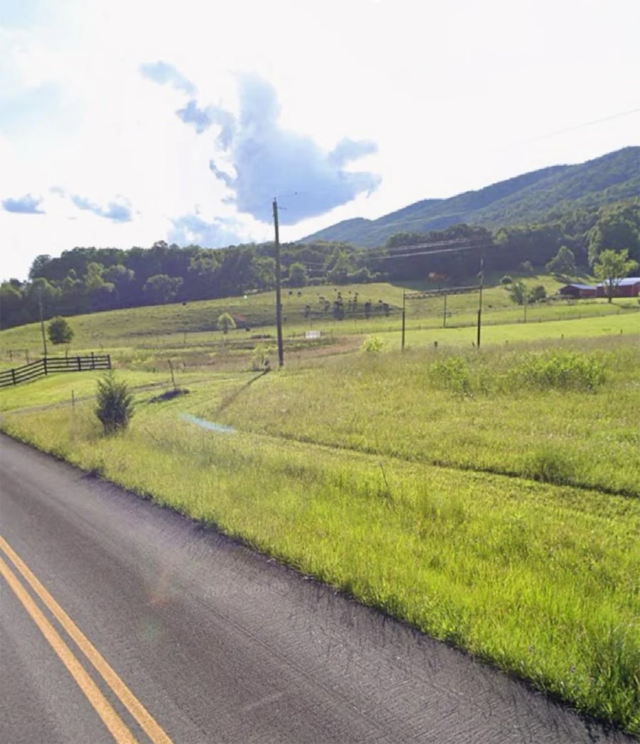 A rural road runs alongside green fields, fenced pastures, and scattered trees, with hills and a partly cloudy sky in the background.