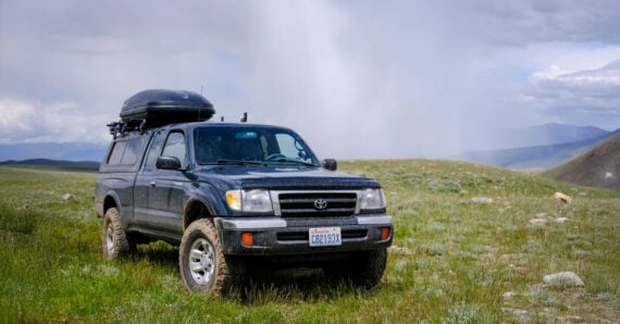 A black Toyota pickup truck with a roof box is parked on a grassy field with mountains and clouds in the background.