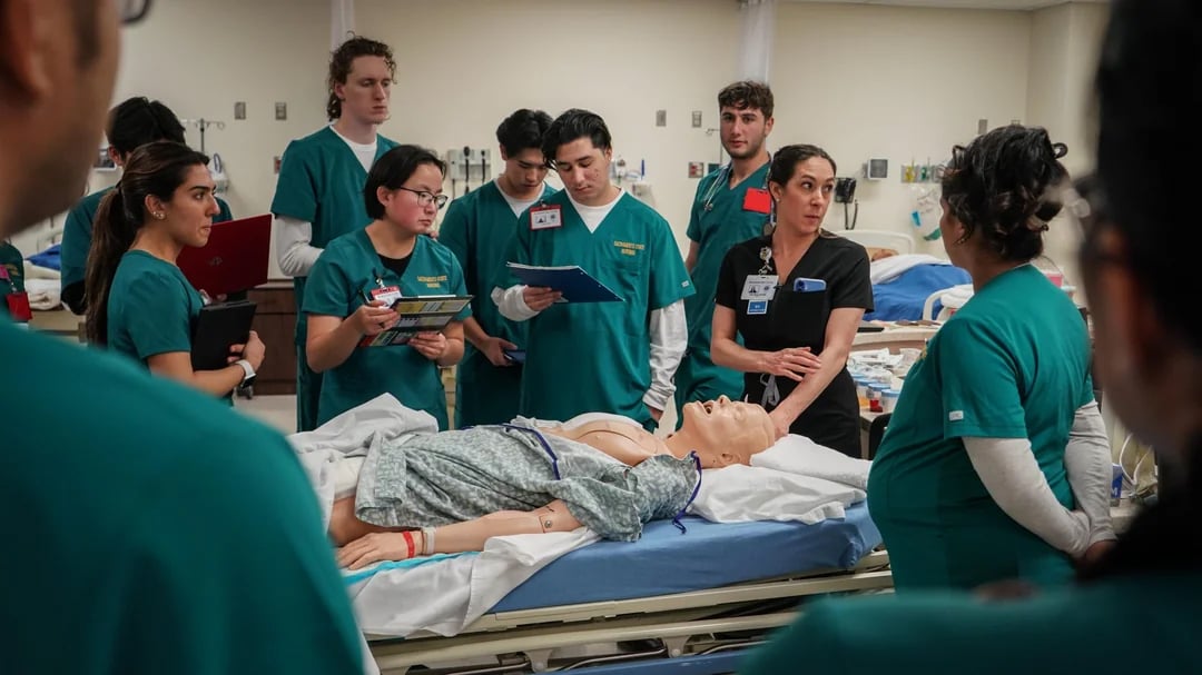 Nursing students in green scrubs gather around a hospital bed with a medical mannequin, taking notes and listening to an instructor in a clinical classroom setting.