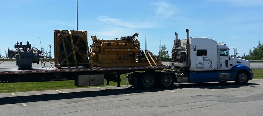 A white and blue semi-truck with a flatbed trailer is parked, transporting large industrial equipment, including a yellow engine and a black machine, in a parking lot on a sunny day.