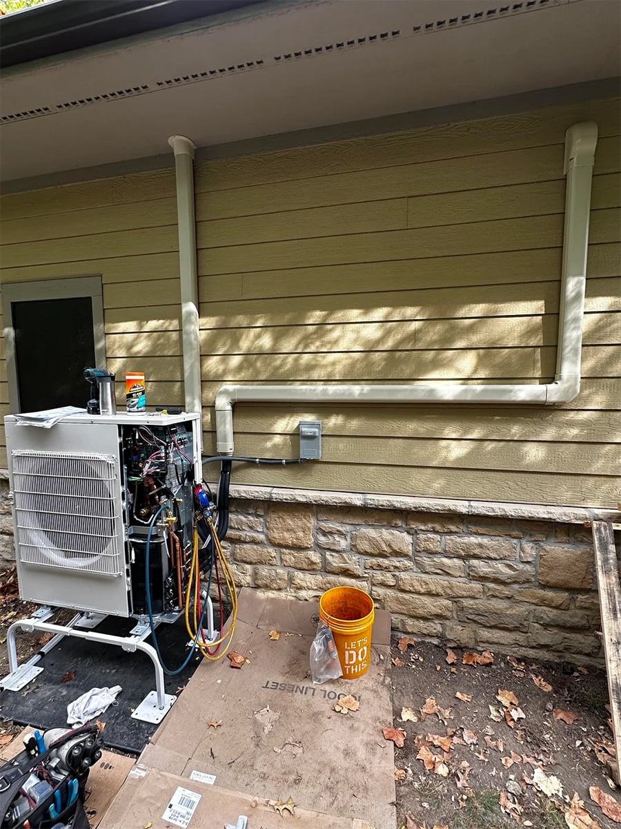 An outdoor HVAC unit with open panels is being serviced near a yellow house wall. Insulated pipes run along the wall. Tools and a yellow bucket are on the ground.