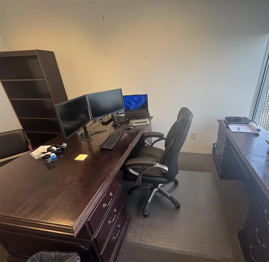 A tidy office with a wooden desk, dual monitors, keyboard, mouse, office chair, empty bookshelf, another desk, scattered electronics, and a window on the right letting in natural light.