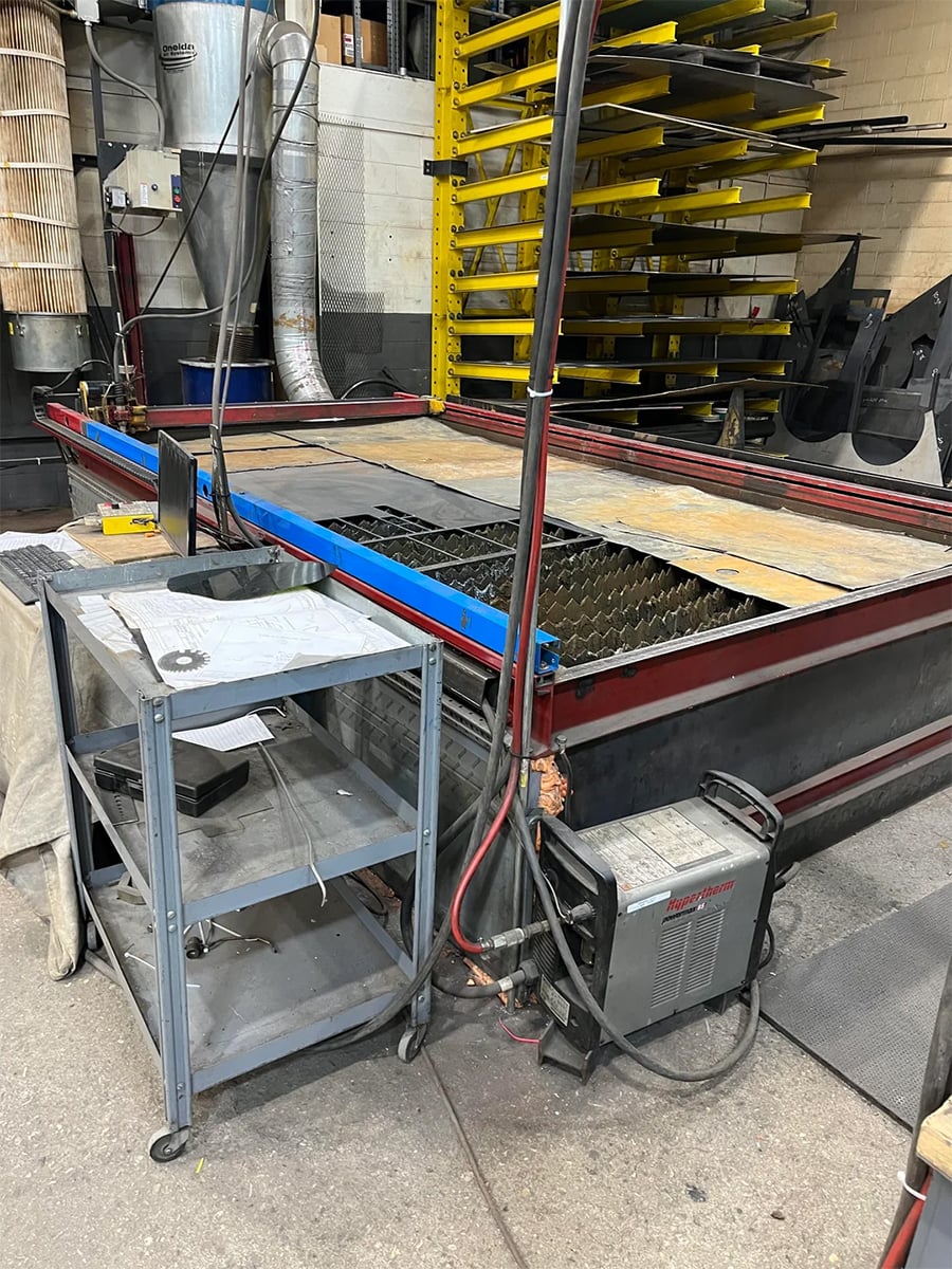 A metal workshop features a CNC plasma cutting table, a welding machine, and a small gray utility cart with tools. Yellow metal shelves and ventilation pipes are in the background.