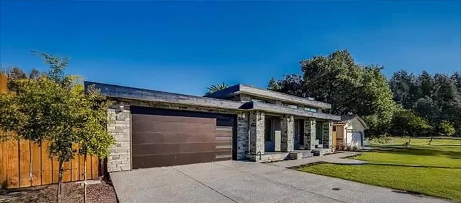 Modern single-story house with a flat roof, stone and wood accents, large dark garage door, and a spacious driveway. Green lawn and trees surround the home under a clear blue sky.