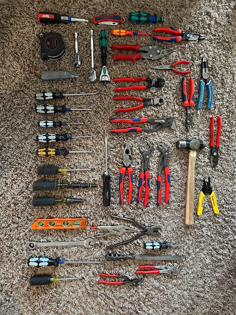 A variety of hand tools including screwdrivers, pliers, wrenches, tape measure, hammer, level, and utility knife arranged neatly on a brown carpet.