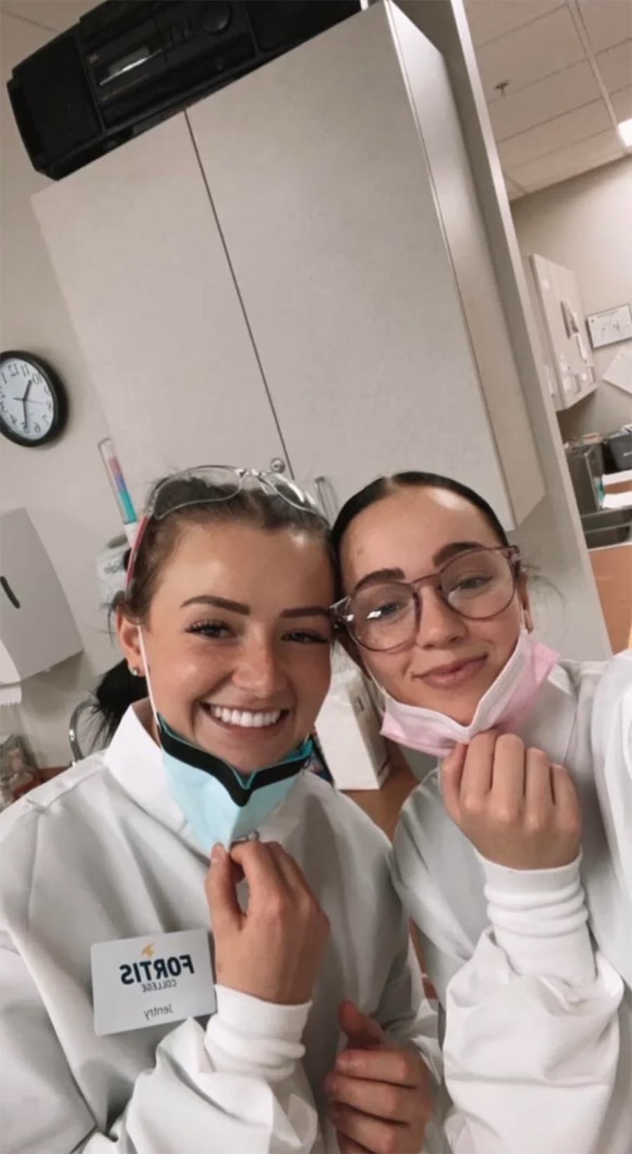 Two women in white lab coats and glasses smile for a selfie in a medical or lab setting. Both are holding their face masks down. A clock and cabinets are visible in the background.