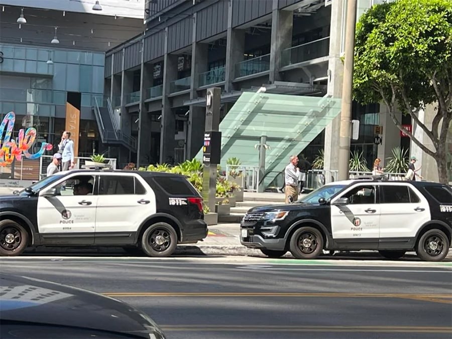 Two black and white police SUVs are parked on a city street near a modern glass building, with a few people walking on the sidewalk in the background.