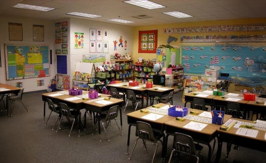 A colorful elementary classroom with student desks arranged in groups, each desk holding papers and supplies. The walls are decorated with educational posters, charts, and a world mural. Shelves hold books and learning materials.