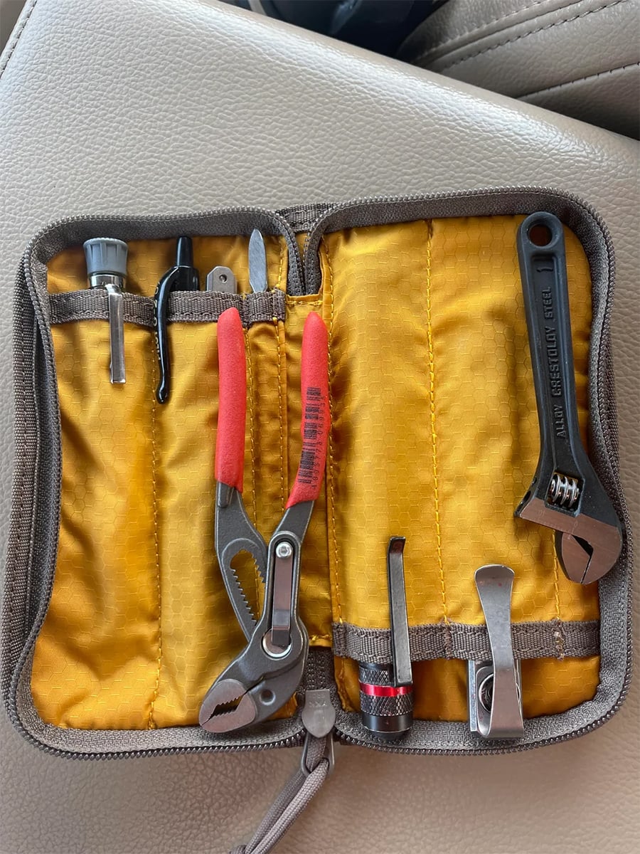 A small yellow tool pouch holding pliers, an adjustable wrench, a screwdriver, two nail clippers, nail scissors, and a small socket, placed on a beige car seat.