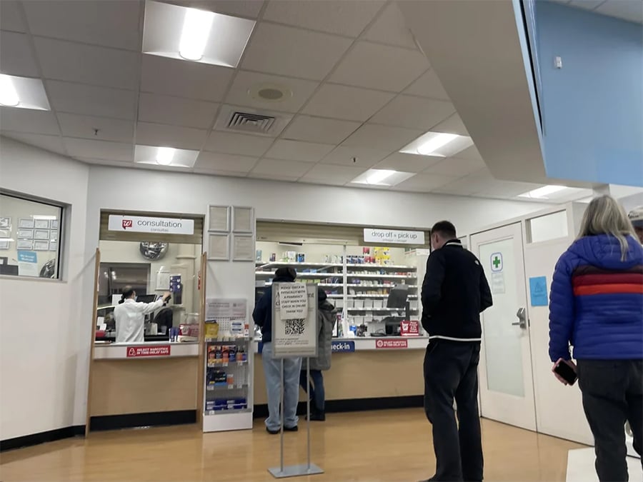 People stand in line at a pharmacy counter inside a clinic. Signs for consultation and prescription pickup are visible. Shelves of medications are seen behind the counter. The floor is wooden, and the lighting is bright.