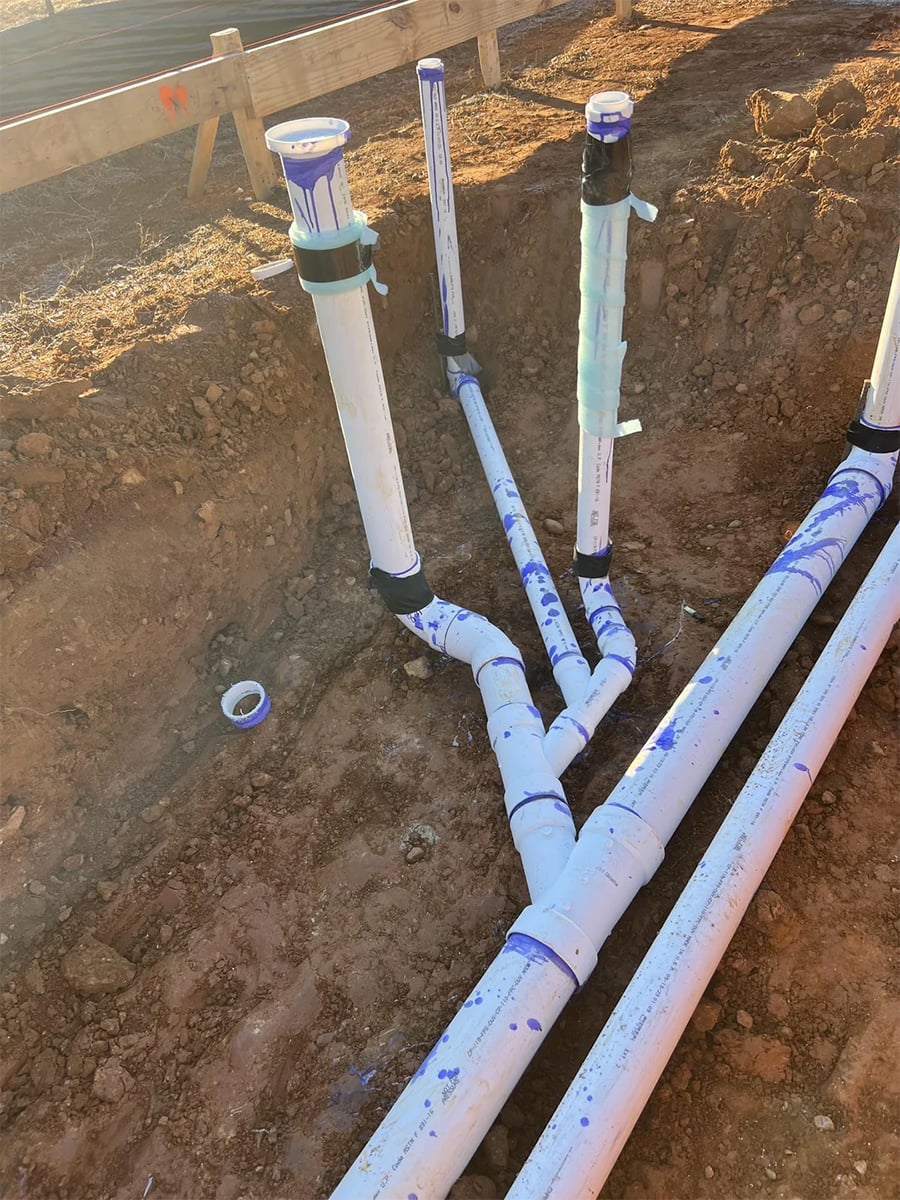 White PVC plumbing pipes are installed in a dirt trench at a construction site. Some pipes are upright, while others are horizontal, and blue markings are visible on the pipes. A roll of tape lies nearby on the ground.